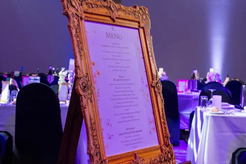 Ornate framed event menu displayed on a table at a masquerade gala dinner. Custom Printed Gala Menus