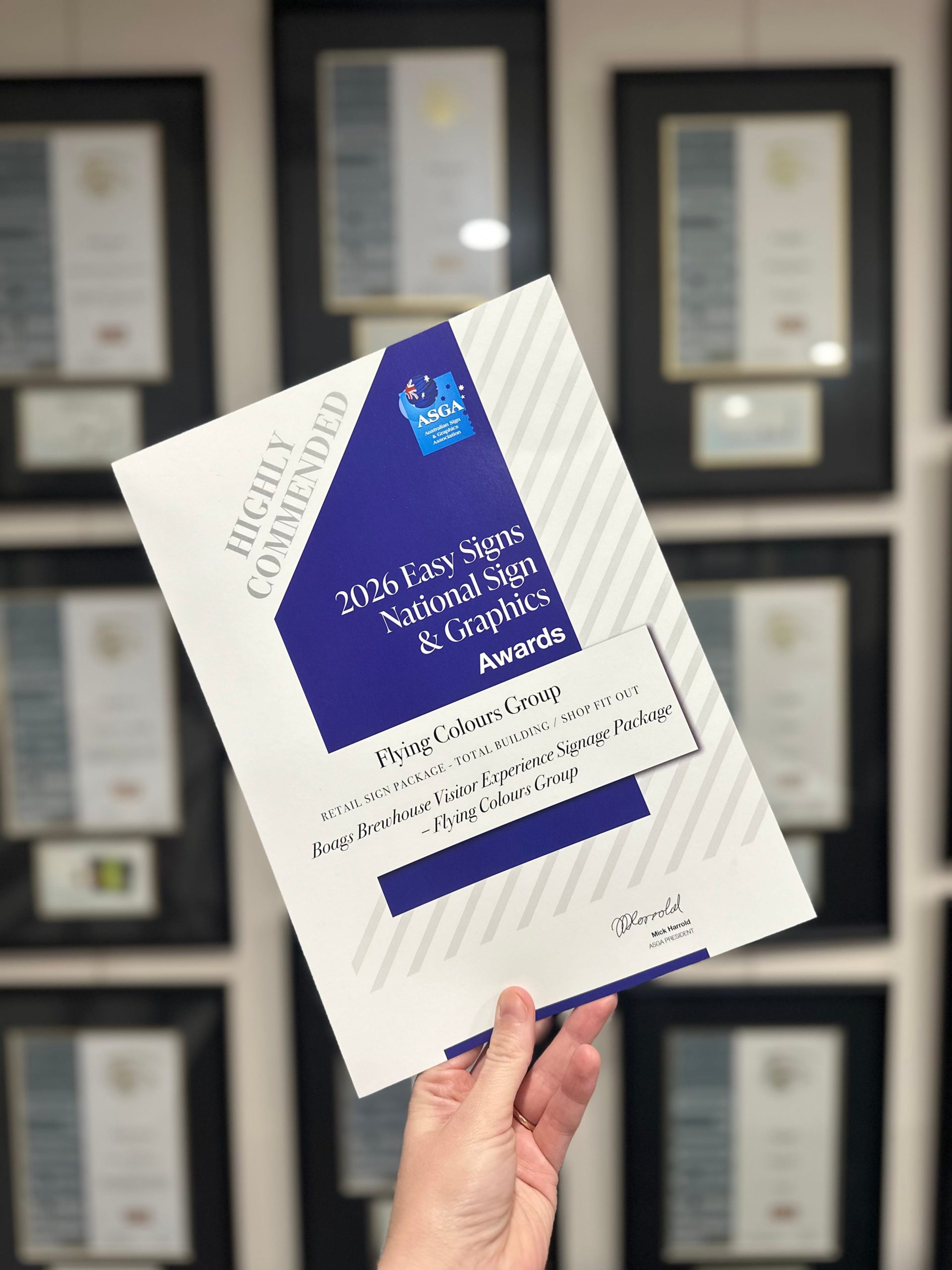Hand holding Flying Colours Group’s 2026 ASGA Highly Commended certificate for Boags Brewhouse signage, with framed awards behind it.