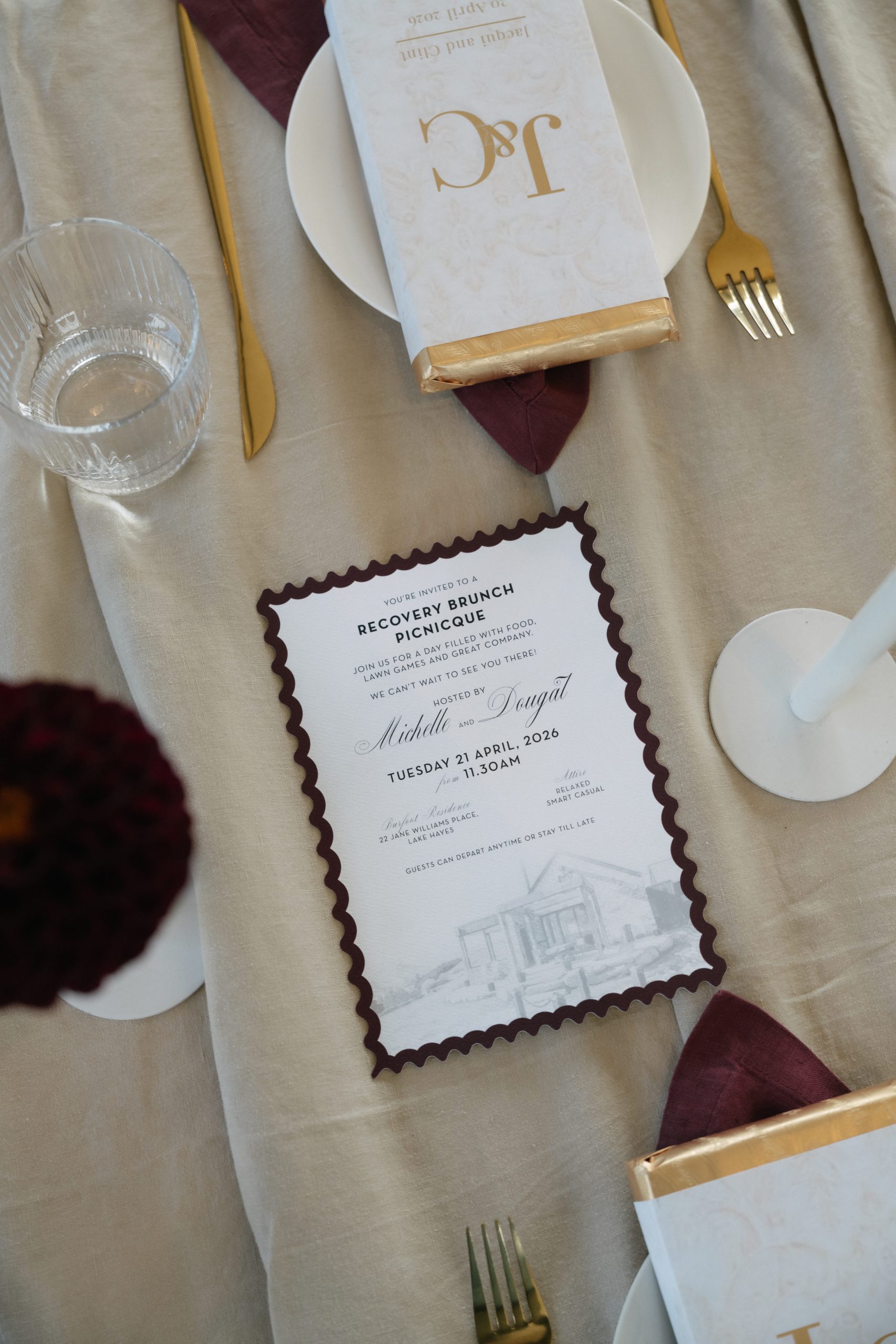 Jacqui and Clint Recovery Brunch Invitation Recovery brunch invitation with burgundy scalloped edges and a house illustration, displayed on ivory linen beside gold cutlery.