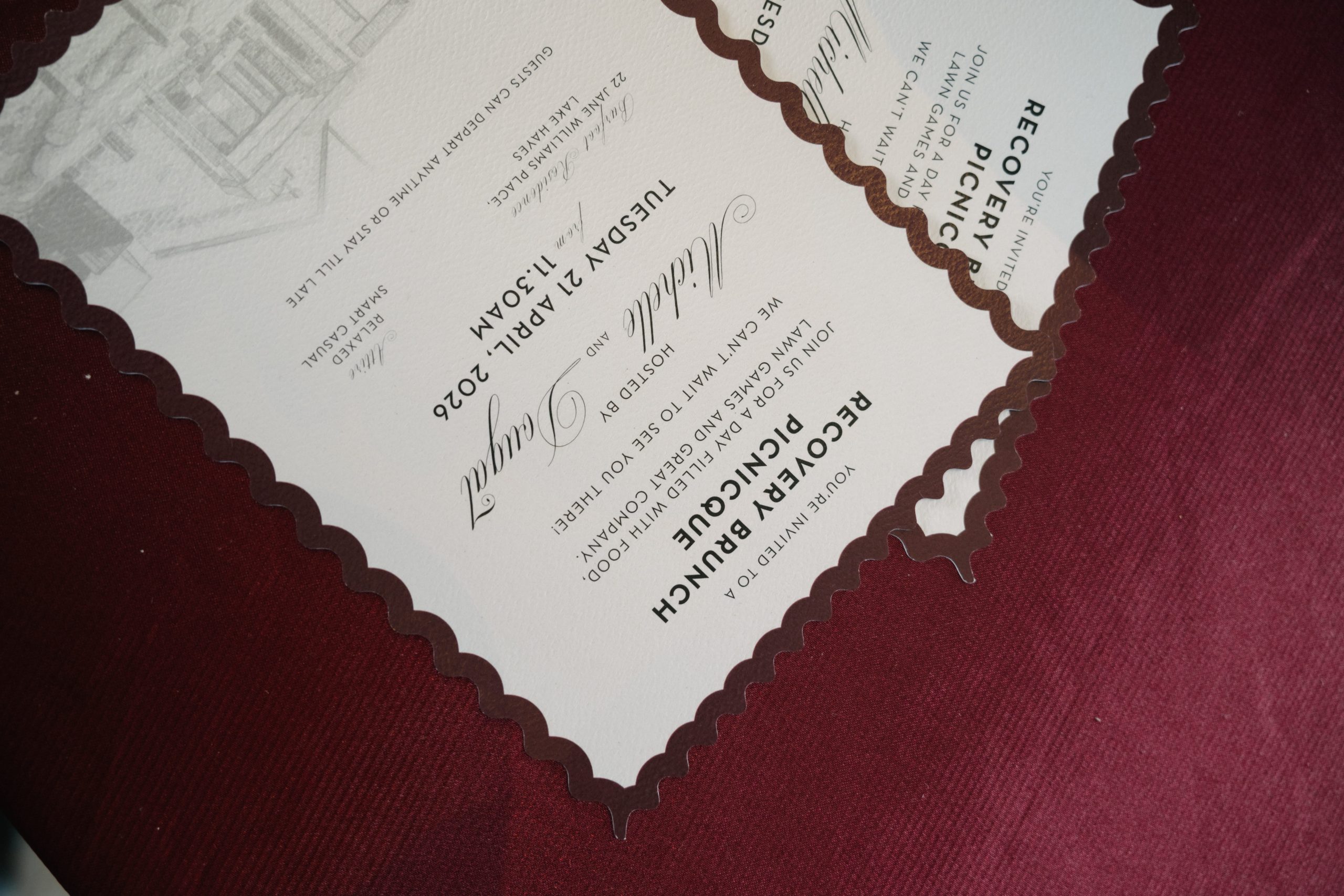Scalloped Recovery Brunch Invitations for Jacqui and Clint Close-up of overlapping ivory recovery brunch invitations with burgundy scalloped borders, script lettering and a house illustration on burgundy fabric.