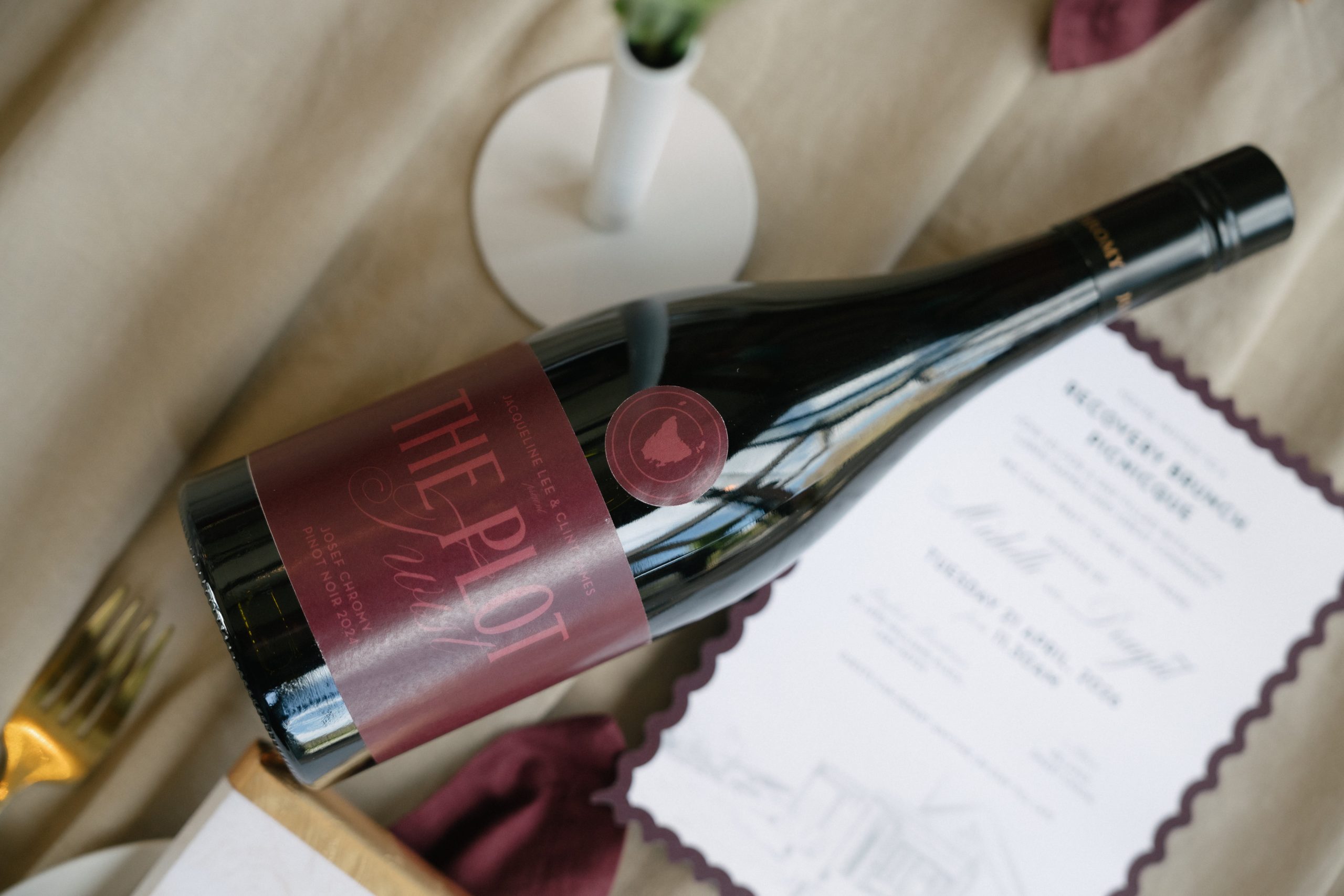 The Plot Twist Custom Wedding Wine Label Wine bottle with a custom burgundy “The Plot Twist” wedding label and round Tasmania sticker beside a scalloped brunch invitation.