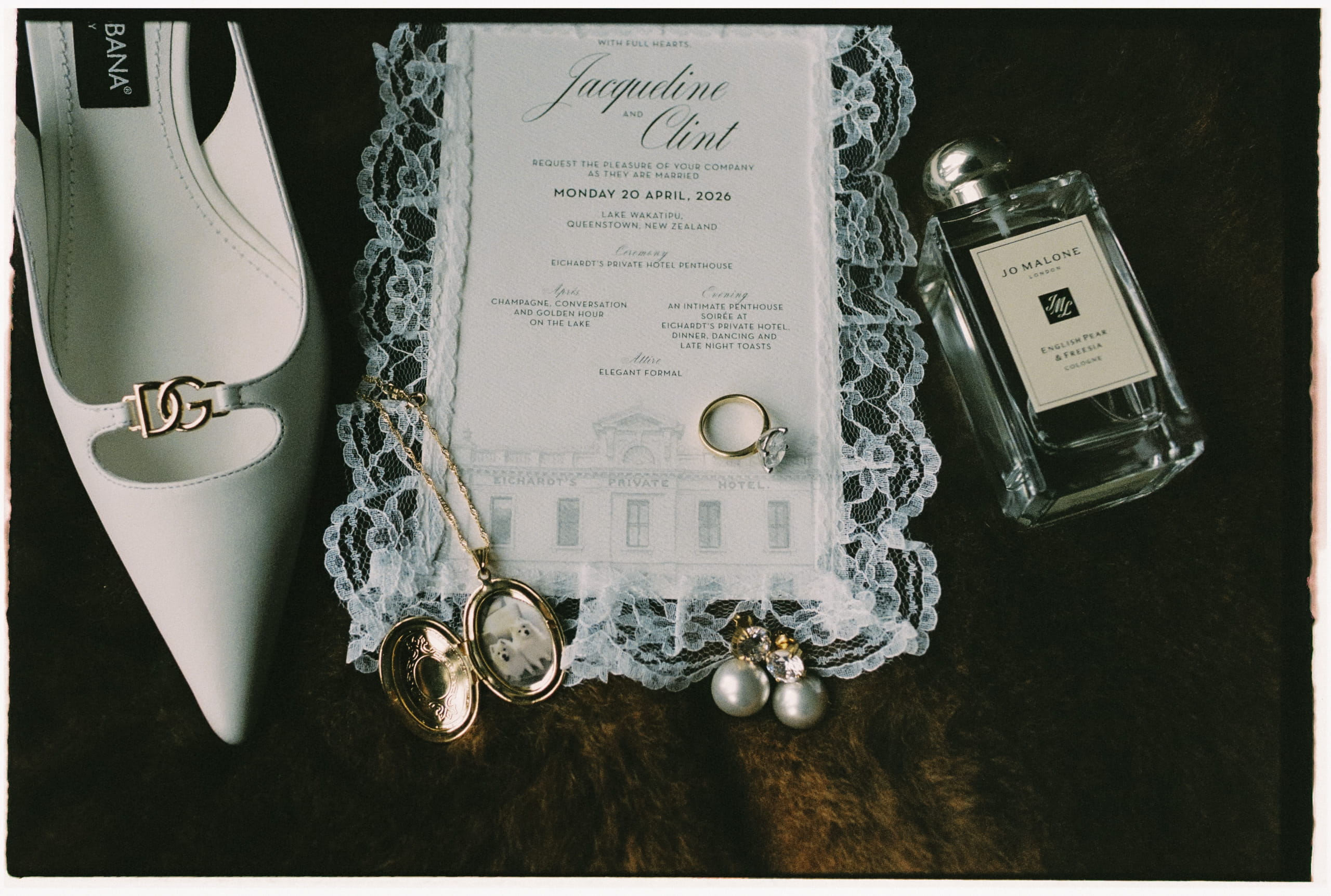 Jacqui and Clint Lace Wedding Invitation Lace-bordered wedding invitation for Jacqueline and Clint with a venue illustration, styled beside a bridal shoe, rings, locket and perfume.
