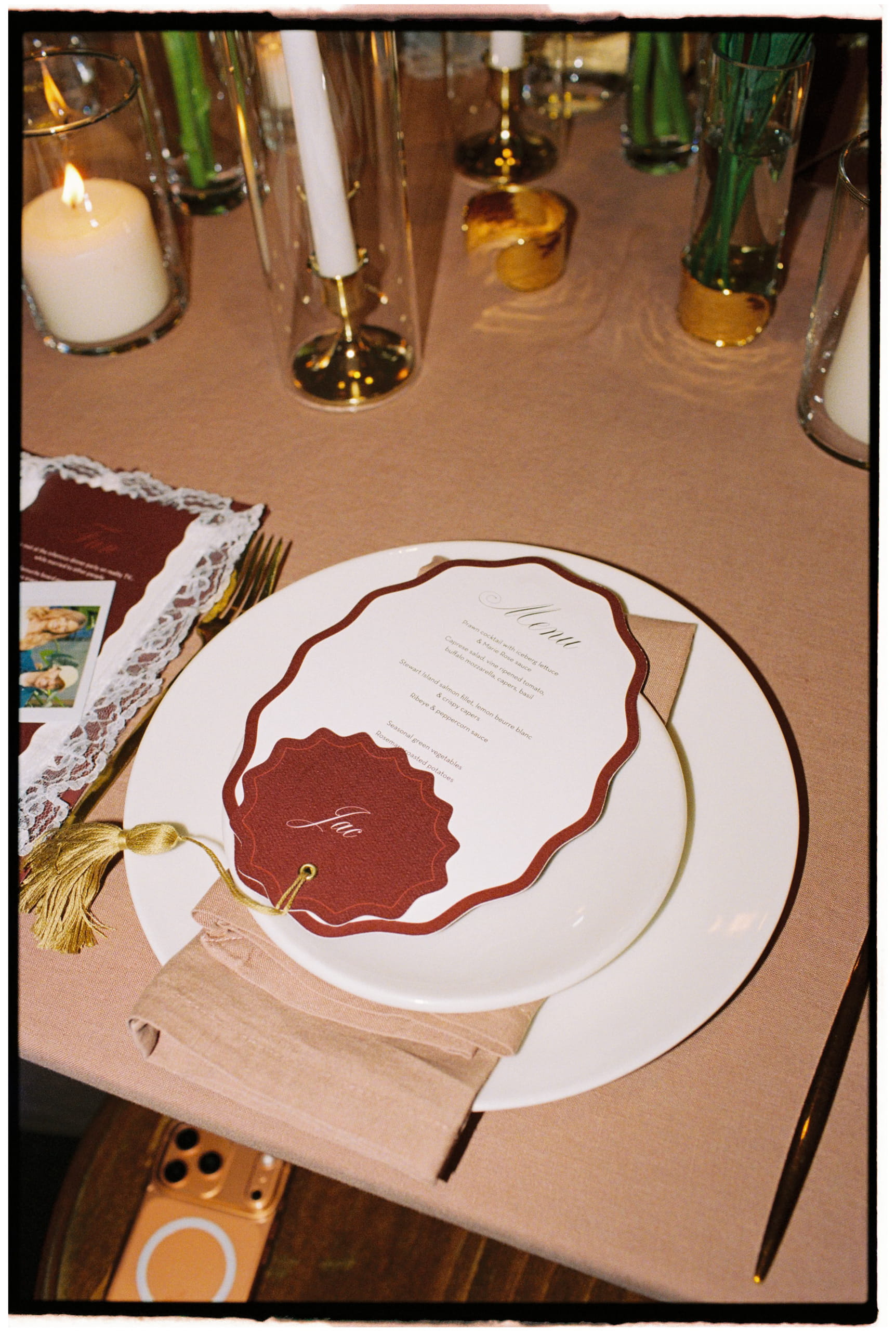 Custom Wedding Menu and Personalised Place Card Ivory wedding menu with a burgundy shaped border and Jac’s personalised gold-tassel name tag on a candlelit reception table.