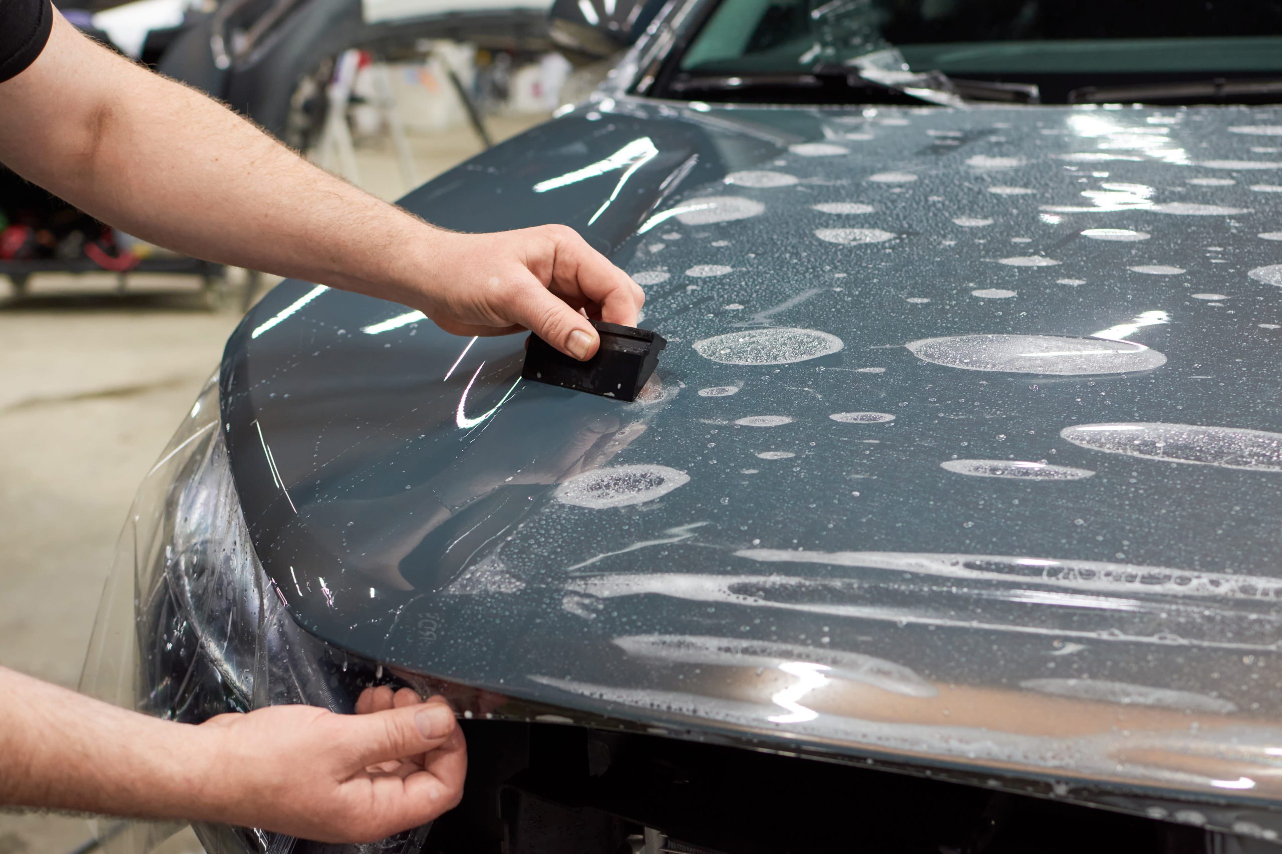 Paint Protection Film Application – Bonnet Installer smoothing clear paint protection film onto a grey vehicle bonnet with a squeegee.